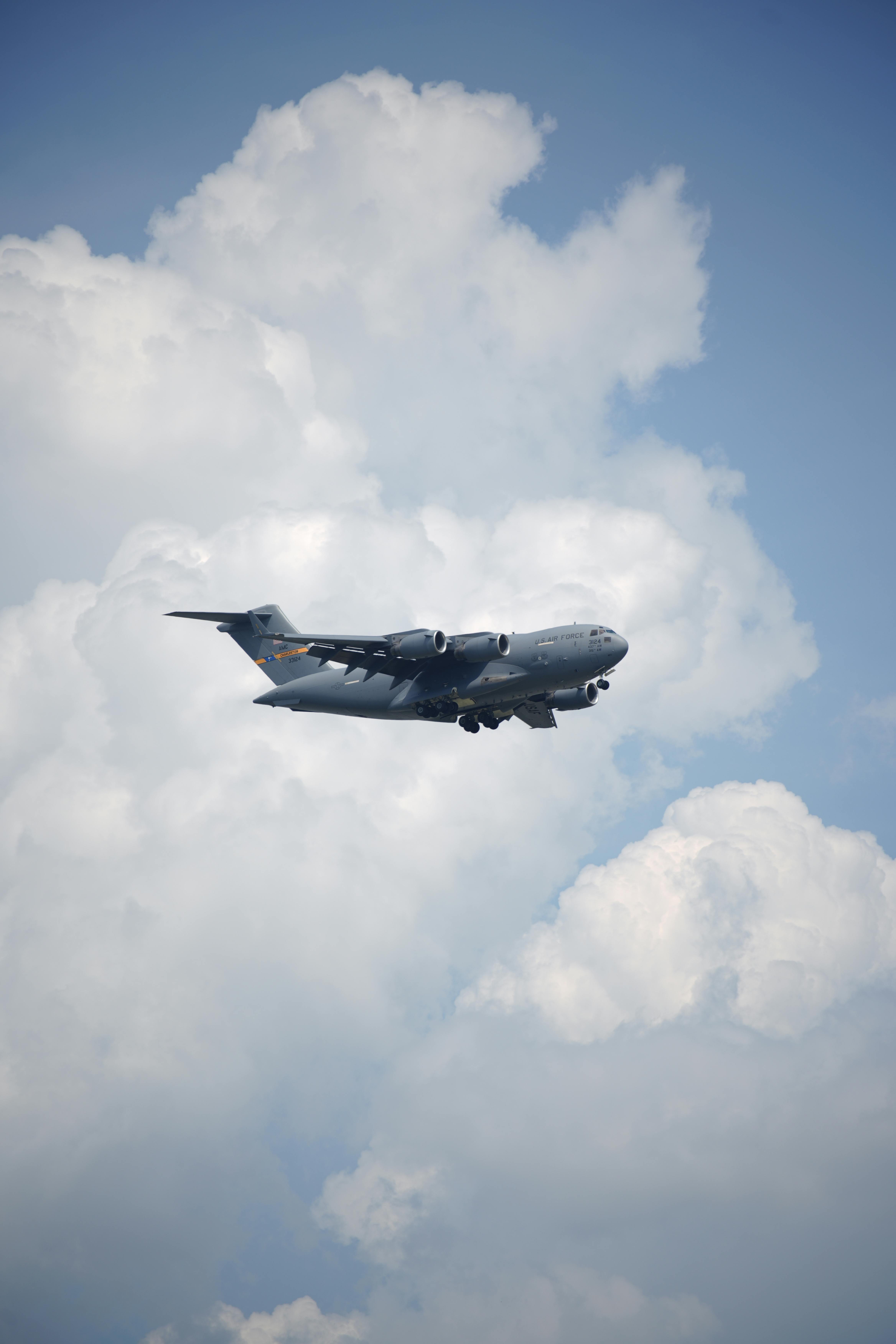 pexels-photo-24339828-24339828 A US Air Force C-17 Globemaster III aircraft flying amidst fluffy clouds in a clear blue sky.